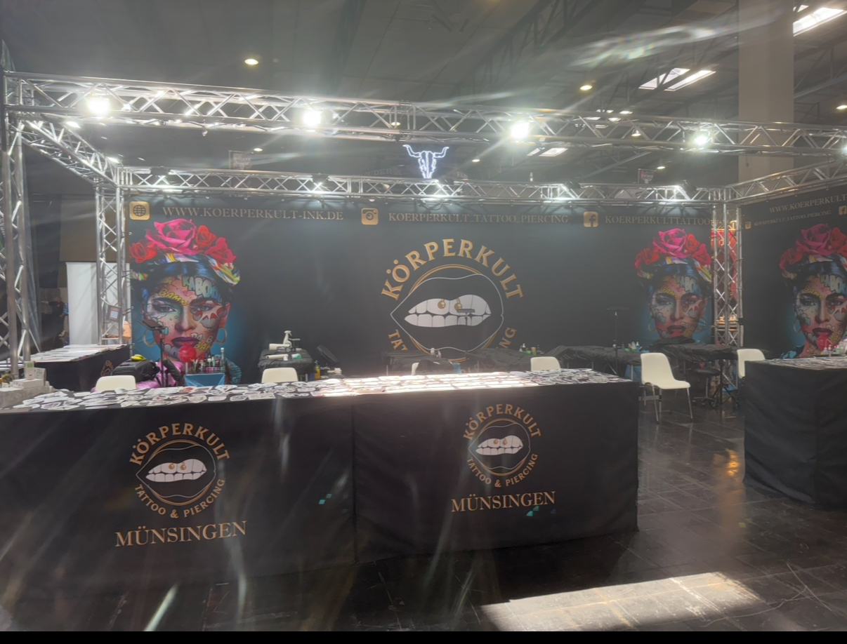 Messestand mit buntem Kunstporträt, Logos und Möbeln. Schwarz-goldenes Farbschema.