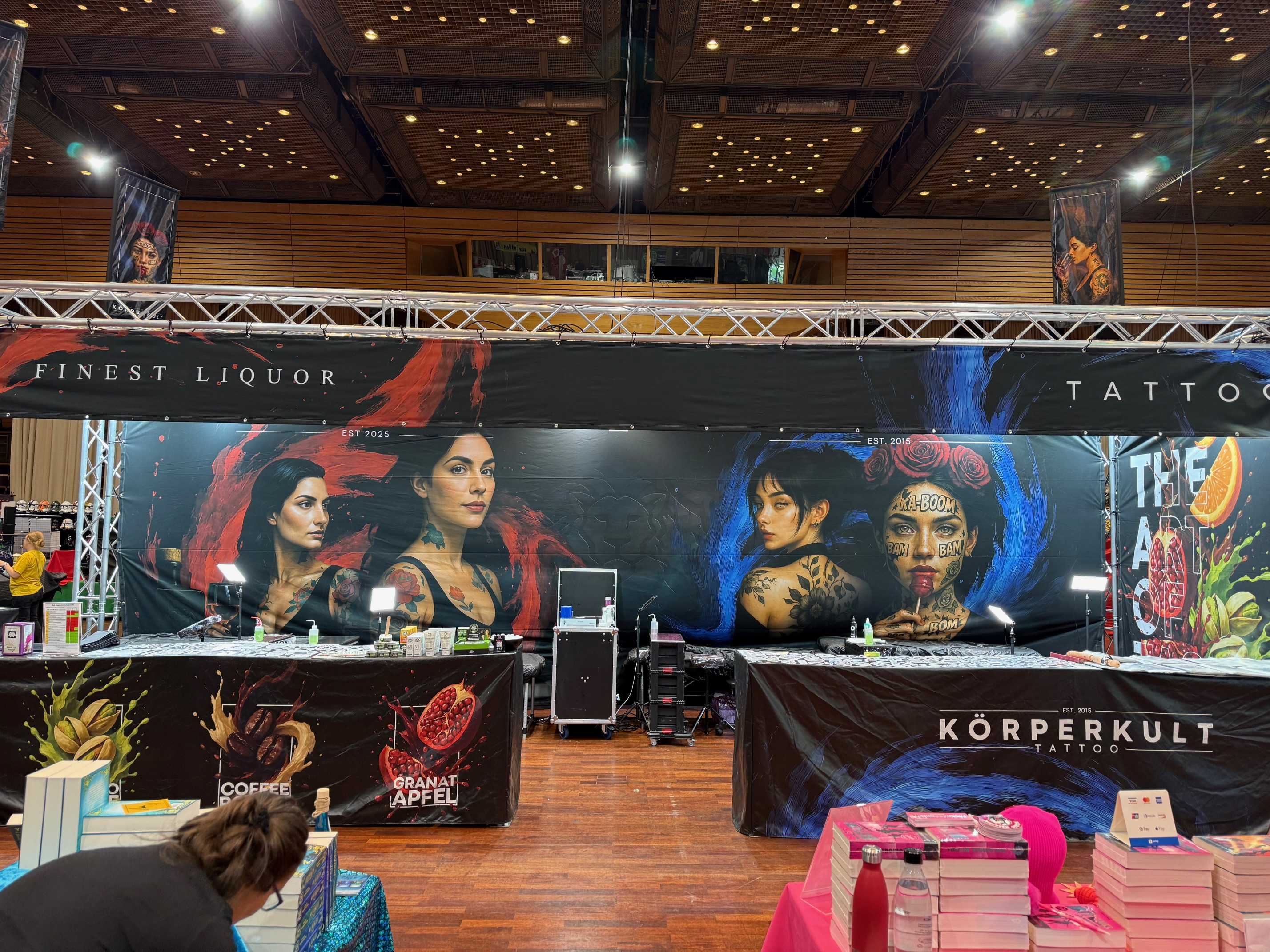 Messehalle mit voller Tattoos und Merchandise vor buntem Wandbild.