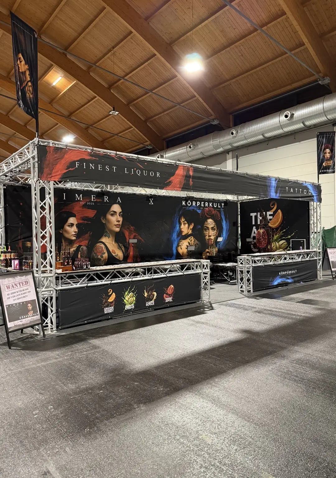 Messestand mit schwarzen Bannern fuer Likoer und Tattoo zeigt taetowierte Frauen